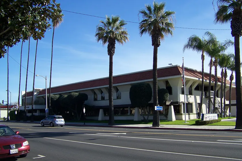 Hemet City Hall, a white two-story building with a red tile roof, set back behind tall palm trees on a sunny street