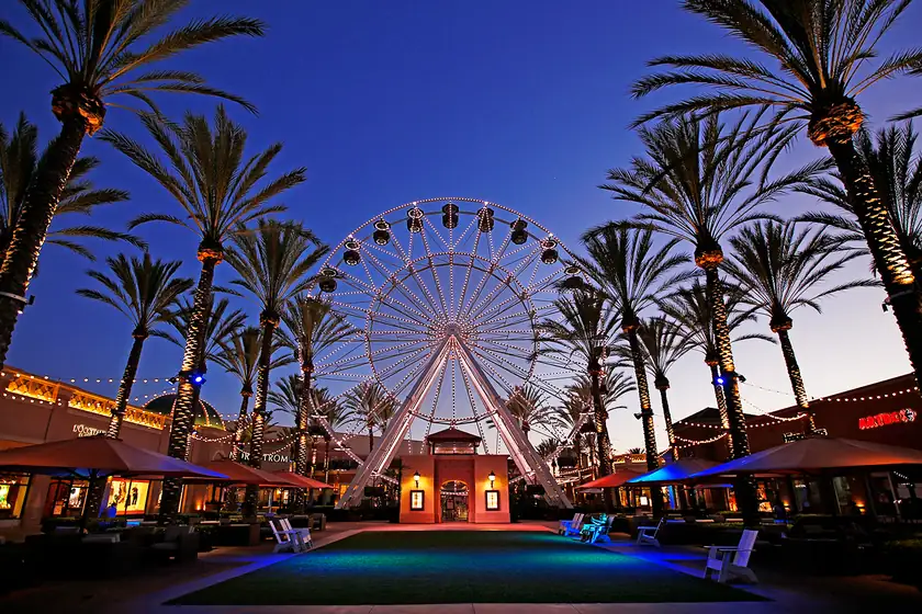 The Giant Wheel at Irvine Spectrum Center lit up at dusk, framed by rows of tall palm trees wrapped in string lights